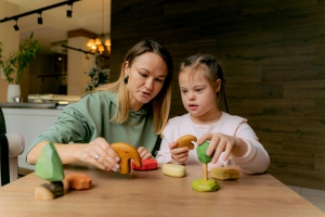 A young girl playing with toy blocks with a female adult who is assumingly the guardian. The adult is talking, and it looks like the young girl is listening whilst playing with the blocks.