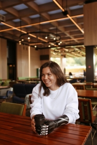 A woman is sat at a table holding a cup of coffee. She is smiling in the photo. Her right arm has a metal prosthetic hand which she is using to hold her cup.