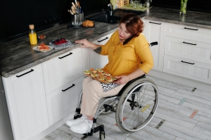 A woman in a wheelchair is cooking in a kitchen. She is also using her mobile phone.