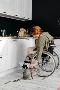 A woman in a wheelchair leaning down to pet a cat. She is in a kitchen.